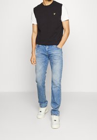 Chaleco negro sin mangas superpuesto sobre una camiseta blanca, combinado con jeans de denim azul claro y zapatillas blancas, mostrando un ajuste relajado.