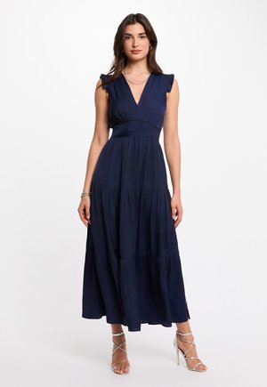 Donna in piedi che indossa un abito lungo senza maniche blu navy con spalle arricciate e sandali con tacco alto argento e cinturini, su sfondo bianco.