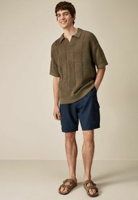 Olive green knitted polo with a textured square pattern, paired with navy cargo shorts and brown sandals. Simple, relaxed fit.