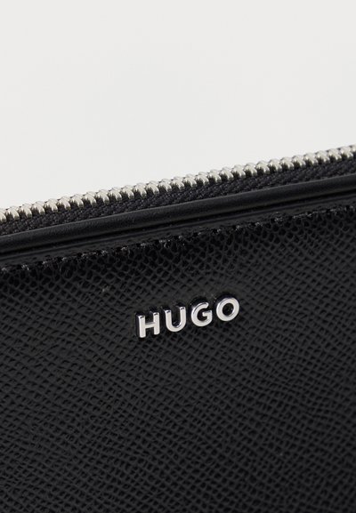 Accessoire en cuir noir texturé avec une fermeture éclair en métal argenté et des lettres argentées formant le mot "HUGO" sur sa surface.