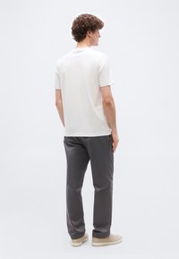 Άνδρας με σγουρά μαλλιά που φοράει άσπρο μπλουζάκι με κοντά μανίκια, γκρι παντελόνι και μπεζ αθλητικά παπούτσια, στέκεται γυρισμένος με την πλάτη σε απλό φόντο.