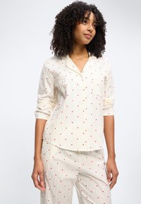 Γυναίκα με σγουρά μαλλιά που φοράει εκρού πιτζάμες με μικρά κόκκινα μοτίβα σε σχήμα καρδιάς, στέκεται σε απλό φωτεινό φόντο.