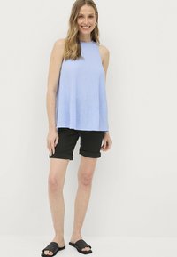 Lichtblauwe mouwloze top met een relaxte pasvorm, gecombineerd met zwarte opgerolde shorts en geweven zwarte slide-sandalen op een witte achtergrond.