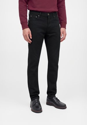 Homme portant un jean slim noir, des chaussures en cuir noir à lacets et une chemise à manches longues rouge foncé sur fond blanc.