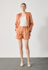 Peach satin blazer and matching shorts, relaxed fit, pleated detail, paired with beige mesh mules, set against a neutral background.