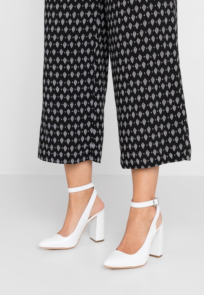Jambes portant un pantalon noir court avec des motifs en losange blancs et des chaussures blanches à talons hauts avec brides à la cheville et bouts pointus.