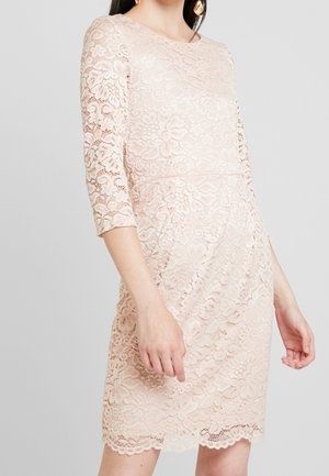 Femme portant une robe en dentelle florale rose pâle avec des manches trois-quarts et un ourlet festonné, visible du cou jusqu'au milieu des cuisses sur un fond gris.