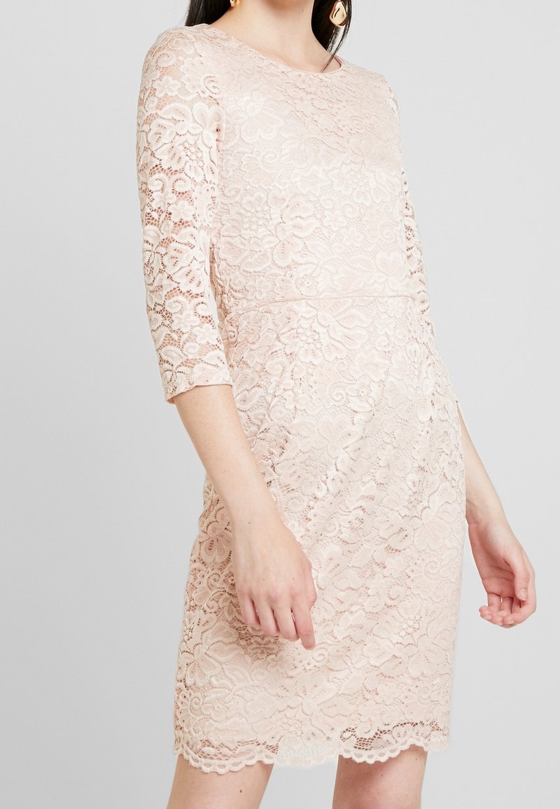 Femme portant une robe en dentelle florale rose pâle avec des manches trois-quarts et un ourlet festonné, visible du cou jusqu'au milieu des cuisses sur un fond gris.