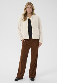 Helles beige Fleece-Cardigan mit Vorderknöpfen, zwei Taschen und lockerer Passform, kombiniert mit braunen cordartigen, weit geschnittenen Hosen und schwarzen Schuhen.