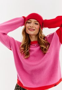 Ružový sveter s červenými akcentmi, rebrovanými manžetami a lemom. Mäkká textúra a voľný strih. Červená pletená čiapka s ohybom.