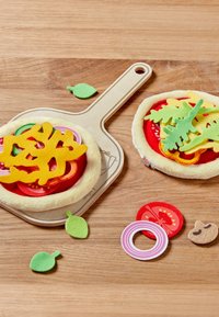 Speelgoed pizza set met vilten toppings in rood, groen, geel en roze op een houten snijplank; inclusief ronde en blaadvormen, met een gepluche basis.