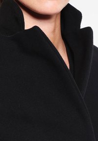 Närbild av en persons nacke och haka iklädd en svart ullkappa med stående krage mot en enfärgad vit bakgrund.