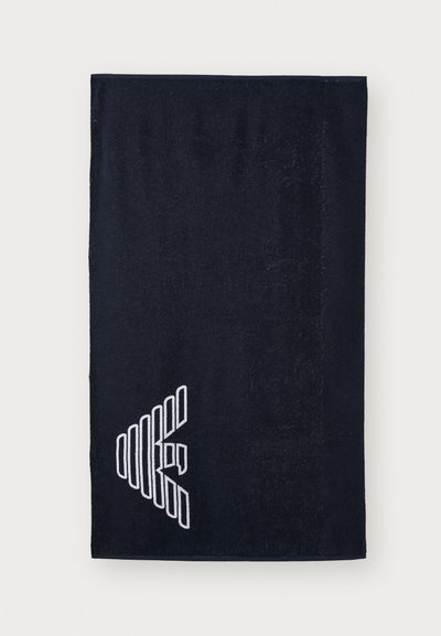 Navy blue towel made of soft material, featuring a white geometric logo at the bottom left. Textured surface with a smooth finish.
