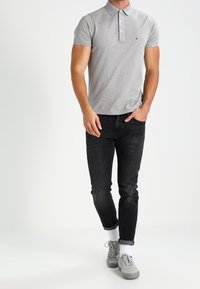Camisa polo gris con cuello, mangas cortas y un pequeño logo. Combinada con jeans ajustados negros y zapatillas gris con calcetines blancos.