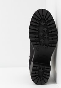 Semelle de botte noire avec un motif de bande de roulement profond, indiquant la taille 37, positionnée verticalement contre un mur d'angle blanc et le sol.