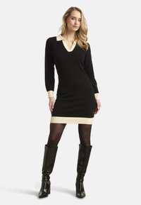 Robe en tricot noir avec col et poignets crème, dotée d'un décolleté en V et d'un ourlet côtelé. Le modèle porte des bottes noires jusqu'aux genoux, avec des collants noirs transparents.