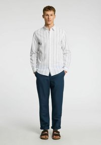 Vit skjorta med marinblå vertikala ränder, tillsammans med mörkblå byxor och bruna sandaler. Outfiten har en avslappnad passform och en avslappnad stil.