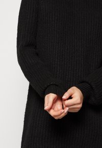 Pull noir en tricot avec un motif texturé. Manches longues avec des poignets côtelés, mains jointes au niveau de la taille.