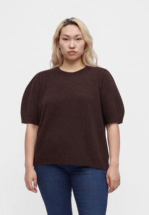 Kvinde med langt blondt hår iført en mørkebrun kortærmet sweater og blå jeans, stående mod en ensfarvet lys baggrund.