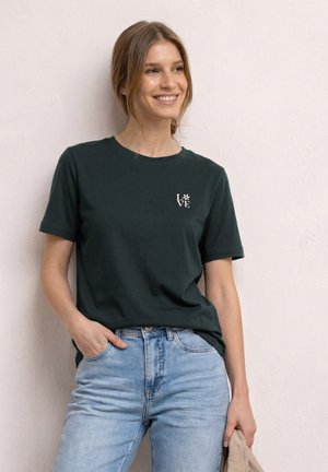 Smilende kvinde iført en mørkegrøn T-shirt med "LOVE" og blomsterdesign, lyseblå jeans, stående op ad en lys tekstureret væg.
