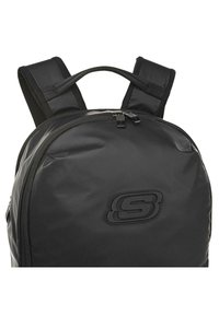 Mochila negra hecha de material duradero, con un diseño redondeado, correas acolchadas y un logo elevado en la parte frontal. Compartimento principal con cremallera.