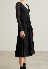 Robes noires avec un corsage côtelé, un décolleté en V et de longues manches translucides. La jupe est fluide et plissée, atteignant la mi-mollet.