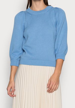 Femme portant un pull en maille bleu clair avec des manches bouffantes trois-quarts et une jupe plissée de couleur crème.