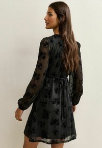 Czarna sukienka w kwiaty z przezroczystymi, długimi rękawami, ściągniętym w pasie i rozkloszowanym dołem. Posiada teksturowane, czarne hafty kwiatowe na lekkim materiale.