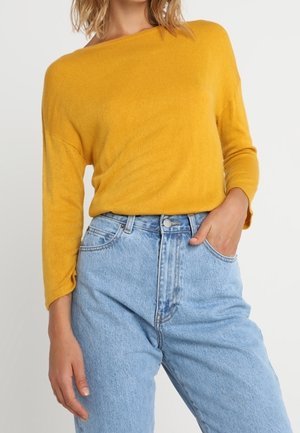 Kvinde iført en karrygul langærmet sweater stoppet ned i højtaljede lyseblå jeans med den ene hånd i lommen.