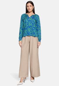 Niebieska bluzka z długim rękawem w zielone wzory liści, zestawiona z beżowymi szerokimi spodniami i białymi szpilkami. Bluzka ma dekolt w kształcie litery V i marszczone rękawy.