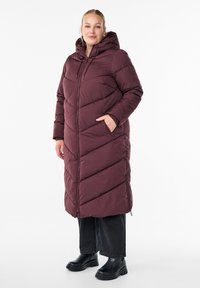 Žena u dugom burgundskom pamučnom kaputu s kapuljačom, crnim širokim hlačama i crnim čizmama na debelu petu, stoji s jednom rukom u džepu.