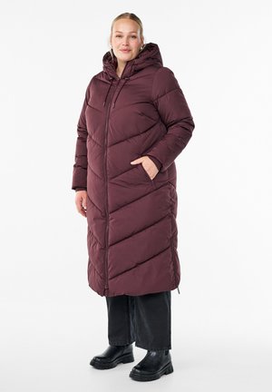 Femme portant un long manteau matelassé bordeaux avec capuche, un pantalon noir à jambes larges et des bottes noires imposantes, se tenant avec une main dans la poche.