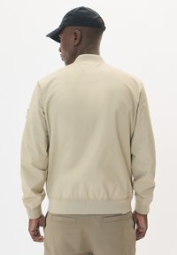 Chaqueta bomber beige con ribetes en el dobladillo y los puños, que presenta una textura suave y mate. Lleva puesta una gorra negra; vista desde la parte de atrás.