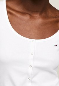 Top de tirantes blanco con canalé que presenta un escote redondo, cierre de botones y un pequeño logo en la zona del pecho. Textura de tela suave.