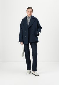 Cappotto di lana doppiopetto blu marina con ampi revers, abbinato a un dolcevita a righe, jeans scuri e scarpe slip-on grigio chiaro. Borsa piccola.