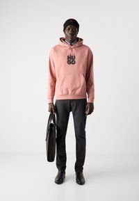 Sudadera con capucha rosa con el logo "HUGO", jeans de mezclilla oscuros, gorro negro y zapatos negros. Sosteniendo una bolsa negra. Diseño sencillo, estilo casual.