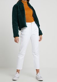 Chaqueta verde de felpa sobre un suéter naranja de cuello alto, combinado con jeans blancos recortados y zapatillas blancas. Texturas suaves y colores contrastantes.