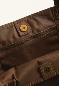 Brown fabric bag interior featuring a dark zipper, golden snap fastener, and embossed brand name "OYSHO." Smooth texture and minimal design.