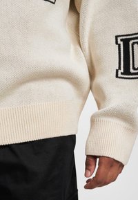 Beige Strickpullover mit gerippten Bündchen und Saum, mit einem schwarzen Logodetail auf der Vorderseite. Die Textur ist weich und leicht.