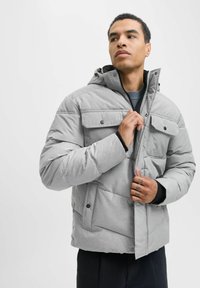 Grå polstret jakke med hætte, der har lynlås lukning, to frontlommer og et struktureret stof. Designet inkluderer trykknap detaljer.