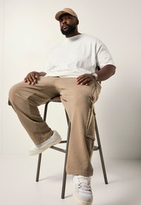 Beige cargobroek en een wit T-shirt, zittend op een kruk. Witte sneakers met grijze accenten maken de outfit af. Een tan cap wordt gedragen.