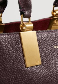 Paarse leren handtas met gouden hardware, voorzien van een rechthoekig label en een textuur oppervlakte. De handvatten hebben een accent van donker leer.