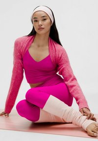 Růžový fitness outfit s crop topem a legínami s vysokým pasem. Měkký růžový kardigan a návleky na nohy ve světle růžové barvě dotvářejí celkový vzhled.