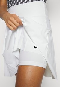 Bílá sportovní sukně s rozšířeným střihem, vrstvenou tkaninou a černým logem Nike. Pod ní jsou vidět bílé přiléhavé šortky. Hladká textura.