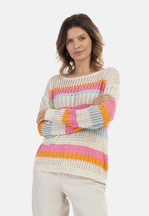 Donna con capelli castani fino alle spalle che indossa un maglione bianco lavorato a maglia con strisce rosa, arancioni e blu, con le braccia incrociate, in piedi davanti a uno sfondo bianco.