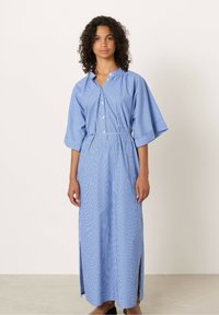 Donna con capelli ricci che indossa un abito maxi a righe blu e bianche con bottoni, maniche ampie e spacco laterale, in piedi contro uno sfondo uniforme.