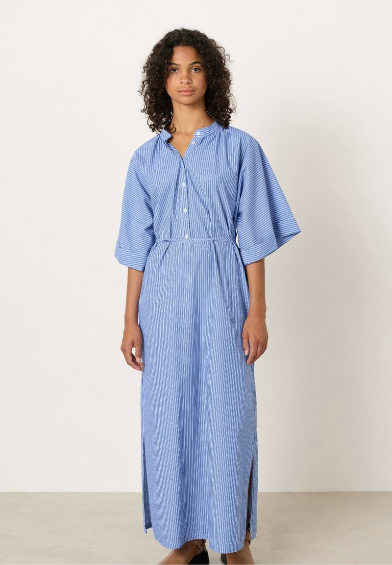 Donna con capelli ricci che indossa un abito maxi a righe blu e bianche con bottoni, maniche ampie e spacco laterale, in piedi contro uno sfondo uniforme.
