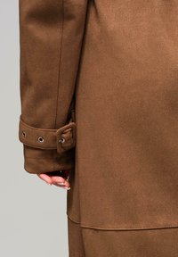 Abrigo marrón de mezcla de lana con un acabado texturizado. Presenta un puño abotonado con ojales metálicos y una silueta elegante.