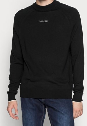 Mężczyzna w czarnym swetrze Calvin Klein i niebieskich dżinsach, pokazany od szyi do bioder, z rękami swobodnie opuszczonymi wzdłuż ciała.