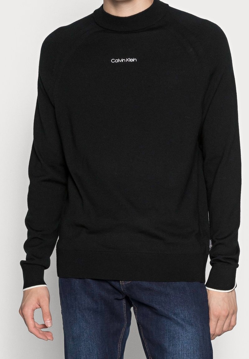 Homme portant un pull noir Calvin Klein et un jean bleu, montré du cou aux hanches avec les mains détendues sur les côtés.